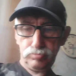 Portret mężczyzny w okularach i czapce z wąsami, z zegarem na ścianie w tle.