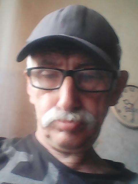 Portret mężczyzny w okularach i czapce z wąsami, z zegarem na ścianie w tle.