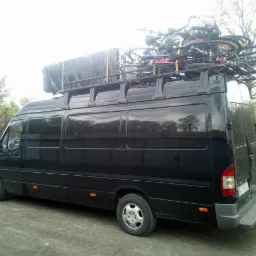 Czarny bus z bagażnikiem dachowym wypełnionym rowerami, zaparkowany na żwirowej drodze, widok z boku i tyłu pojazdu.