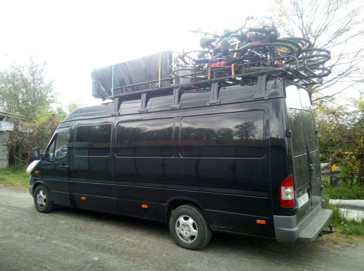 Czarny bus z bagażnikiem dachowym wypełnionym rowerami, zaparkowany na żwirowej drodze, widok z boku i tyłu pojazdu.