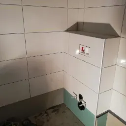 Ściana łazienki w trakcie układania płytek ceramicznych w odcieniach bieli, z widoczną instalacją podtynkową WC i fragmentem ściany wykończonej płytą gipsowo-kartonową, przygotowaną pod dalsze prace.