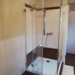 Kabinowa szklana zabudowa prysznicowa z brodzikiem, wykończona brązowymi płytkami, zamontowana w łazience z kremowymi ścianami i listwą dekoracyjną.