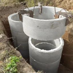 Montaż betonowych kręgów szamba w wykopie, widoczne metalowe uchwyty i łączenia, ziemia i trawa w tle.