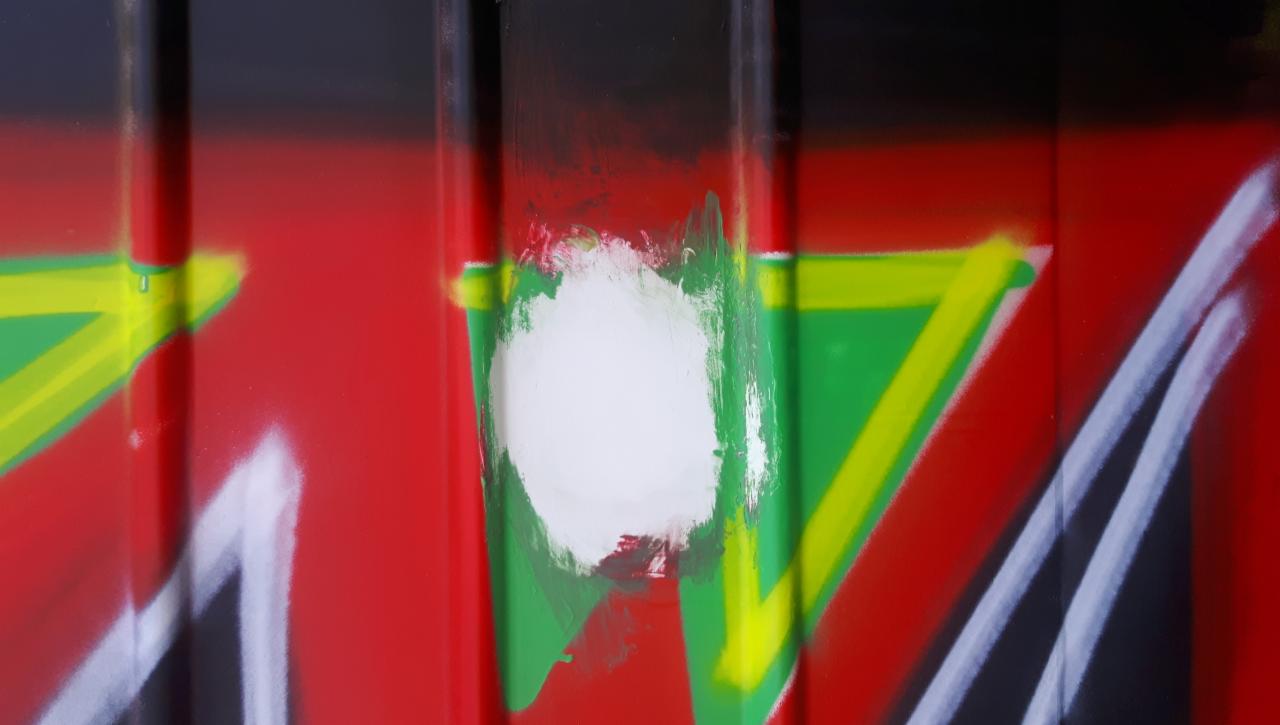 Fragment pomalowanej metalowej powierzchni z widocznymi śladami po usuwaniu graffiti, gdzie biała farba częściowo zakrywa kolorowe elementy.
