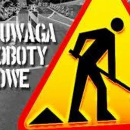 Żółty znak ostrzegawczy z symbolem robotnika z łopatą i napisem 'UWAGA ROBOTY DROGOWE' na tle drogi w Nowym Aleksandrowie, z widocznymi słupkami wygradzającymi teren robót.