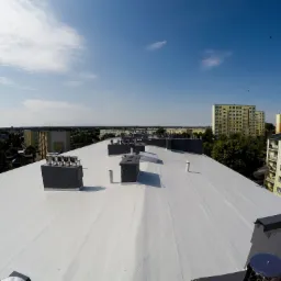 Biały dach po renowacji z kominami i odpowietrznikami na tle miejskiego krajobrazu w słoneczny dzień.