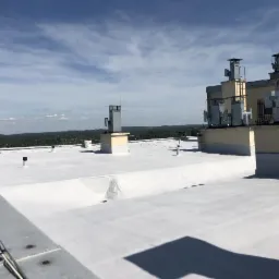 Biała, płaska powierzchnia dachu przemysłowego z kominami wentylacyjnymi i instalacjami, widoczna z góry w słoneczny dzień, z zielonym horyzontem w tle.