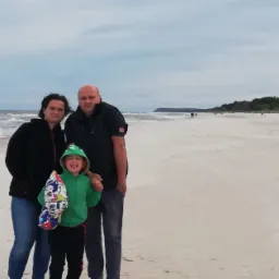 Rodzinne zdjęcie na plaży z szumiącym morzem w tle. Uśmiechnięta dziewczynka w zielonej bluzie i rodzice stoją blisko siebie. Pochmurne niebo nad piaszczystym brzegiem.
