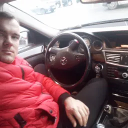 Mężczyzna w czerwonej kurtce siedzi za kierownicą Mercedesa. Widoczna deska rozdzielcza, kierownica z logo i fragment wnętrza samochodu.