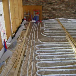 Instalacja ogrzewania podłogowego w trakcie montażu: białe rurki ułożone na srebrnej folii izolacyjnej, widoczne drewniane legary, ściana z cegły i rozdzielacz z rurkami czerwonymi i niebieskimi.