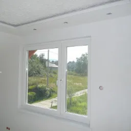 Białe, dwuskrzydłowe okno PCV w nowo wykończonym pokoju z gładkimi, białymi ścianami i ozdobnym sufitem. Widok na zewnątrz ukazuje zielony krajobraz z domami w oddali.