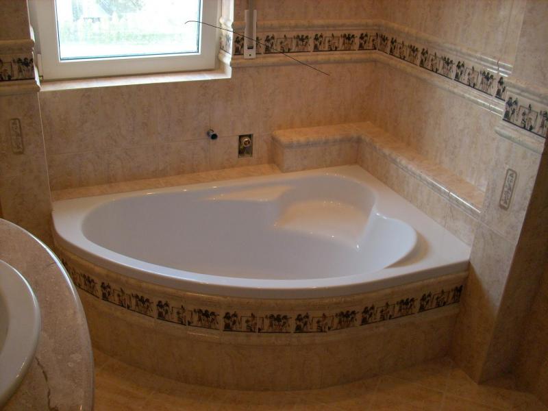 Narożna wanna z hydromasażem w beżowej łazience z dekoracyjnymi płytkami, widok z góry, przy oknie zamontowany biały grzejnik.