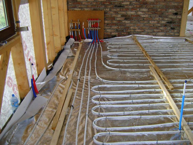 Instalacja ogrzewania podłogowego w trakcie montażu: białe rurki ułożone na srebrnej folii izolacyjnej, widoczne drewniane legary, ściana z cegły i rozdzielacz z rurkami czerwonymi i niebieskimi.