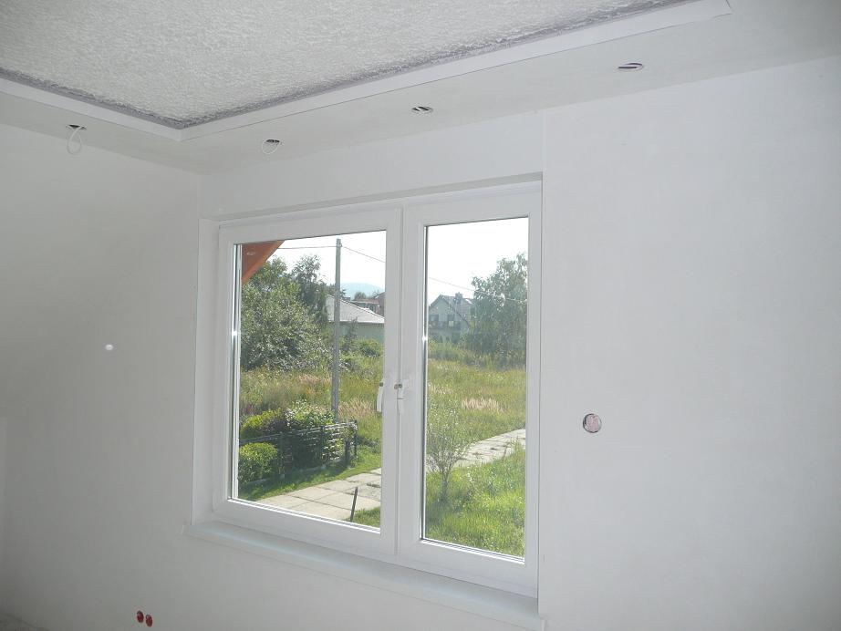 Białe, dwuskrzydłowe okno PCV w nowo wykończonym pokoju z gładkimi, białymi ścianami i ozdobnym sufitem. Widok na zewnątrz ukazuje zielony krajobraz z domami w oddali.