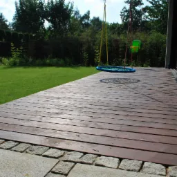 Drewniany taras z desek w odcieniach brązu, obok trawnik i huśtawki na tle zieleni, wykończenie z kamiennej kostki.