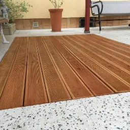 Ułożona drewniana podłoga tarasowa z ryflowanych desek na jasnym, wzorzystym podłożu, widok z góry, w tle rośliny w donicach i biała ławka.