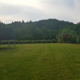 Rozległy trawnik z trampoliną w rogu, w tle drzewa i wzgórze, równo skoszona trawa.