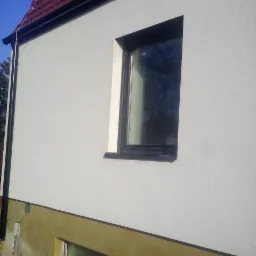 Ściana domu po tynkowaniu na biało, widoczne okno z czarną ramą i fragment ocieplonej, ale jeszcze nieotynkowanej części elewacji przy gruncie.