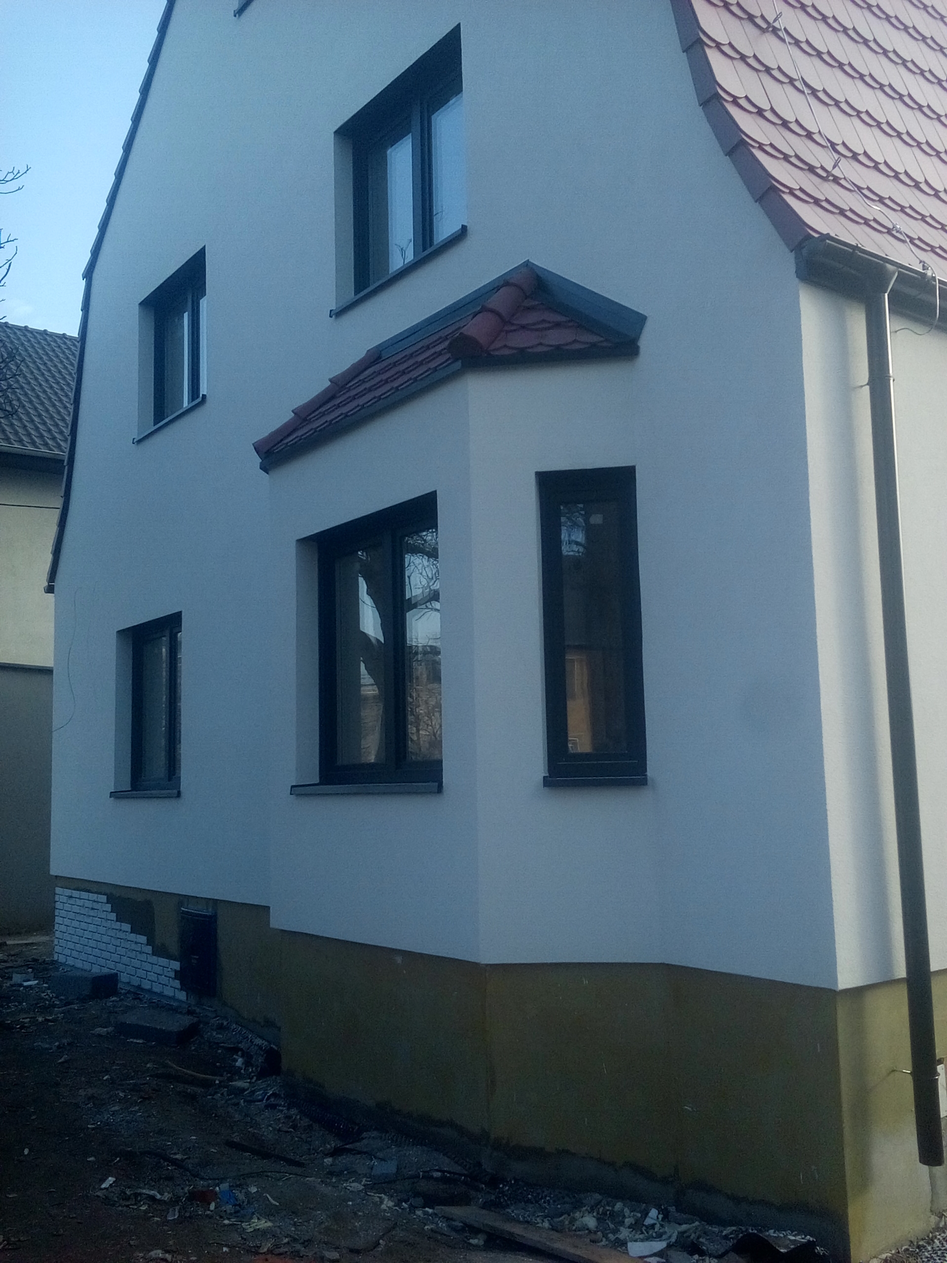 Dwupiętrowy dom z nową, jasną elewacją, czarnymi ramami okiennymi i czerwoną dachówką; widoczne niedokończone prace przy fundamentach i pozostałości materiałów budowlanych.