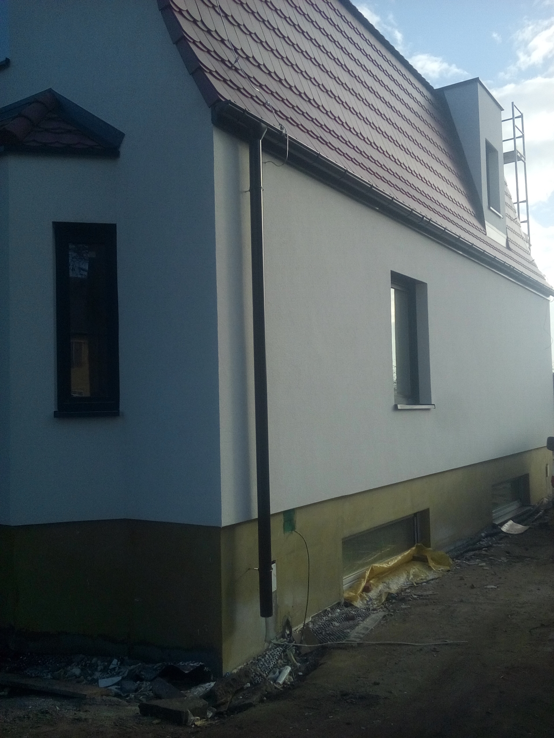 Świeżo otynkowana elewacja domu jednorodzinnego z widocznymi oknami, rynnami i fragmentem dachu z czerwonej dachówki, teren wokół budynku w trakcie prac wykończeniowych.