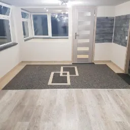 Wykończone wnętrze poddasza z oknami, drzwiami i panelami ściennymi, z dwoma rodzajami podłóg: jasnymi panelami i ciemną podłogą z wzorem geometrycznym.