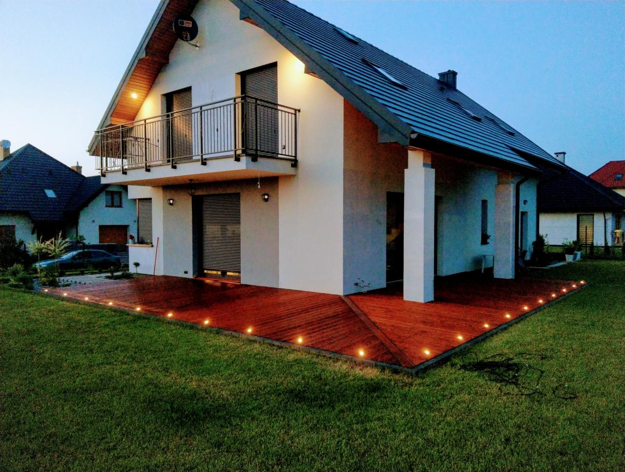 Nowoczesny dom z tarasem z desek oświetlonym punktowymi lampami, widok z ogrodu o zmierzchu.