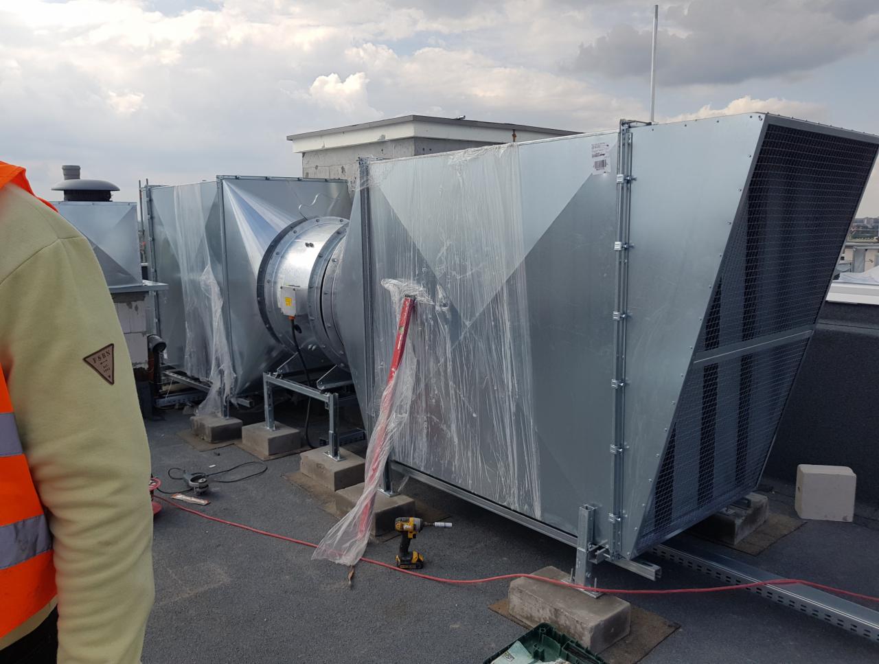 System wentylacyjny na dachu budynku, częściowo owinięty folią ochronną, widoczny fragment osoby w kamizelce odblaskowej oraz narzędzia, pochmurne niebo w tle.