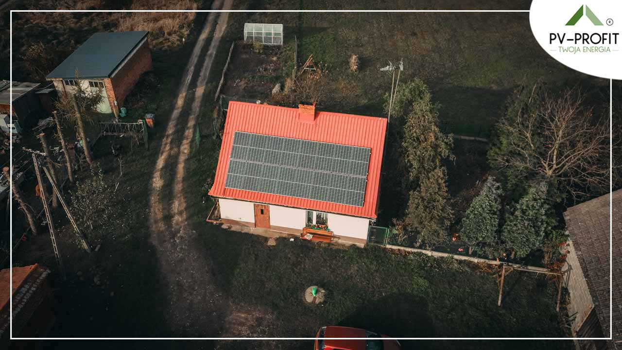 Dom jednorodzinny z czerwonym dachem i panelami fotowoltaicznymi, widziany z lotu ptaka, otoczony drzewami i zabudowaniami gospodarczymi, w tle logo firmy PV-PROFIT.