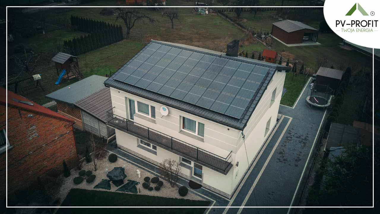 Widok z góry na dom jednorodzinny z zainstalowanymi panelami słonecznymi na dachu, widoczny ogród z elementami zabaw dla dzieci, ciemna kostka brukowa na podjeździe i logo firmy w prawym górnym rogu.