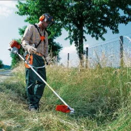 Pracownik w okularach ochronnych i słuchawkach, używa podkaszarki spalinowej do przycinania wysokiej trawy wzdłuż ogrodzenia z siatki.