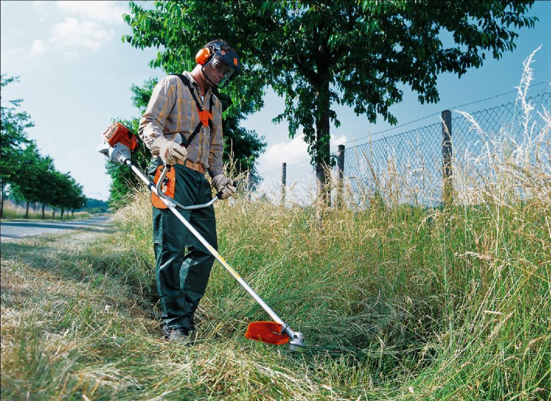 Pracownik w okularach ochronnych i słuchawkach, używa podkaszarki spalinowej do przycinania wysokiej trawy wzdłuż ogrodzenia z siatki.
