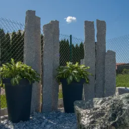 Aranżacja ogrodu z pionowymi, nieregularnymi słupami z szarego kamienia, donicami z roślinami i ozdobnym kamieniem z wodą, na tle siatki ogrodzeniowej i zieleni.