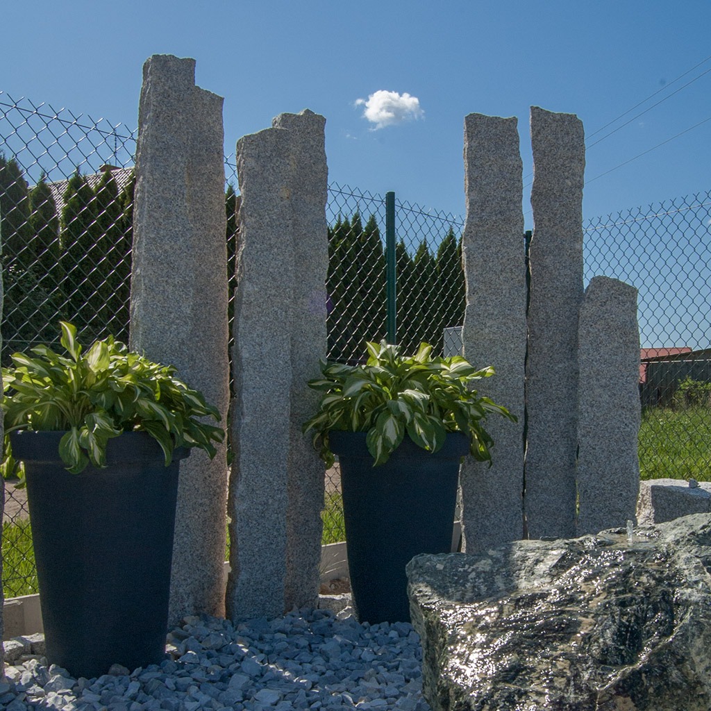 Aranżacja ogrodu z pionowymi, nieregularnymi słupami z szarego kamienia, donicami z roślinami i ozdobnym kamieniem z wodą, na tle siatki ogrodzeniowej i zieleni.