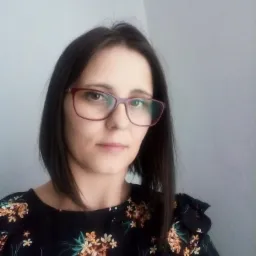 Portret kobiety w okularach i kwiecistej bluzce na tle szarej ściany.