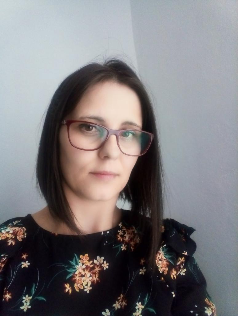 Portret kobiety w okularach i kwiecistej bluzce na tle szarej ściany.