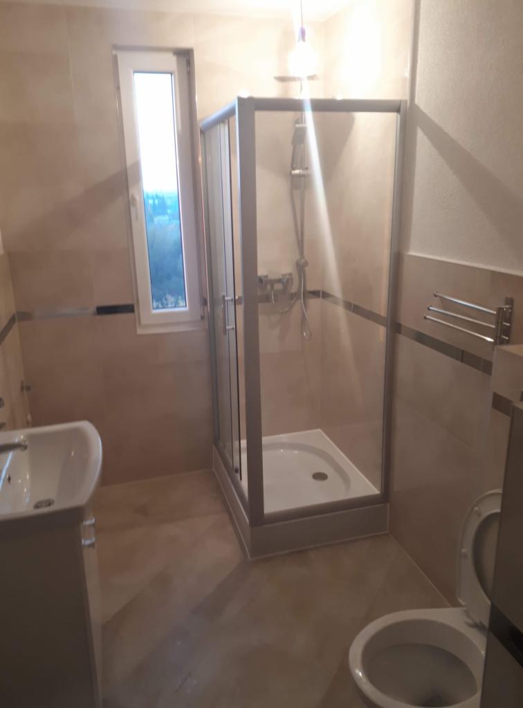 Wykończona łazienka z kabiną prysznicową ze srebrnymi ramami, umywalką na postumencie i toaletą, wykończona w beżowych płytkach z ciemniejszym paskiem dekoracyjnym.