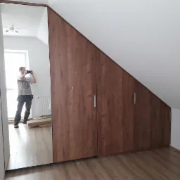 Zabudowa drewniana pod skosem na poddaszu, widoczna na zdjęciu i odbiciu w lustrzanych drzwiach szafy, z metalowymi uchwytami i podłogą imitującą drewno.