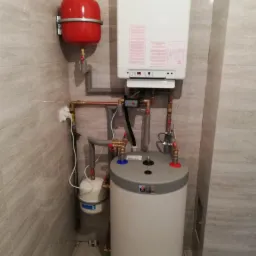 Kompaktowa instalacja grzewcza w łazience z płytkami imitującymi kamień, obejmująca biały kocioł gazowy, czerwony zbiornik wyrównawczy, szary zasobnik ciepłej wody z niebiesko-czerwonymi detalami...