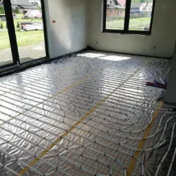 Rozłożona instalacja ogrzewania podłogowego na folii izolacyjnej z widocznymi rurkami i taśmami mocującymi, w surowym stanie wykończenia wnętrza z dużymi oknami.