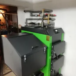 Zielono-czarny piec na paliwo stałe z szarymi rurami instalacji grzewczej w kotłowni, widok z dołu. Nowoczesny model z panelem sterowania.