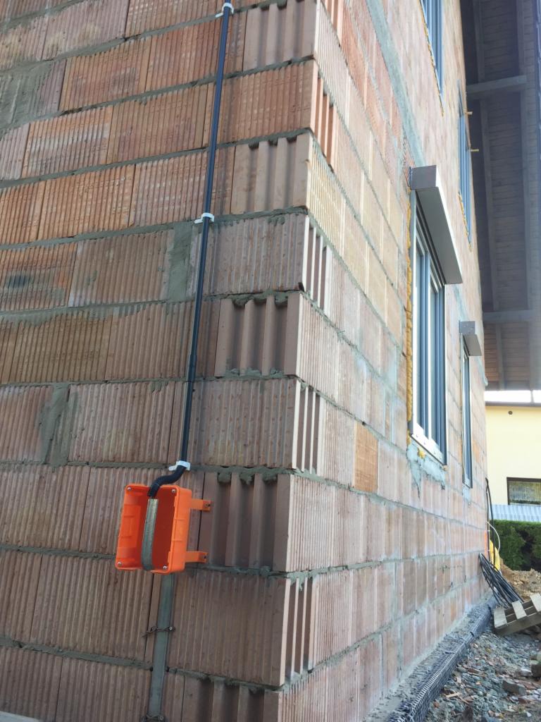 Pomarańczowa puszka elektryczna natynkowa z doprowadzonym przewodem, zamocowana na ścianie z bloczków ceramicznych podczas budowy domu.