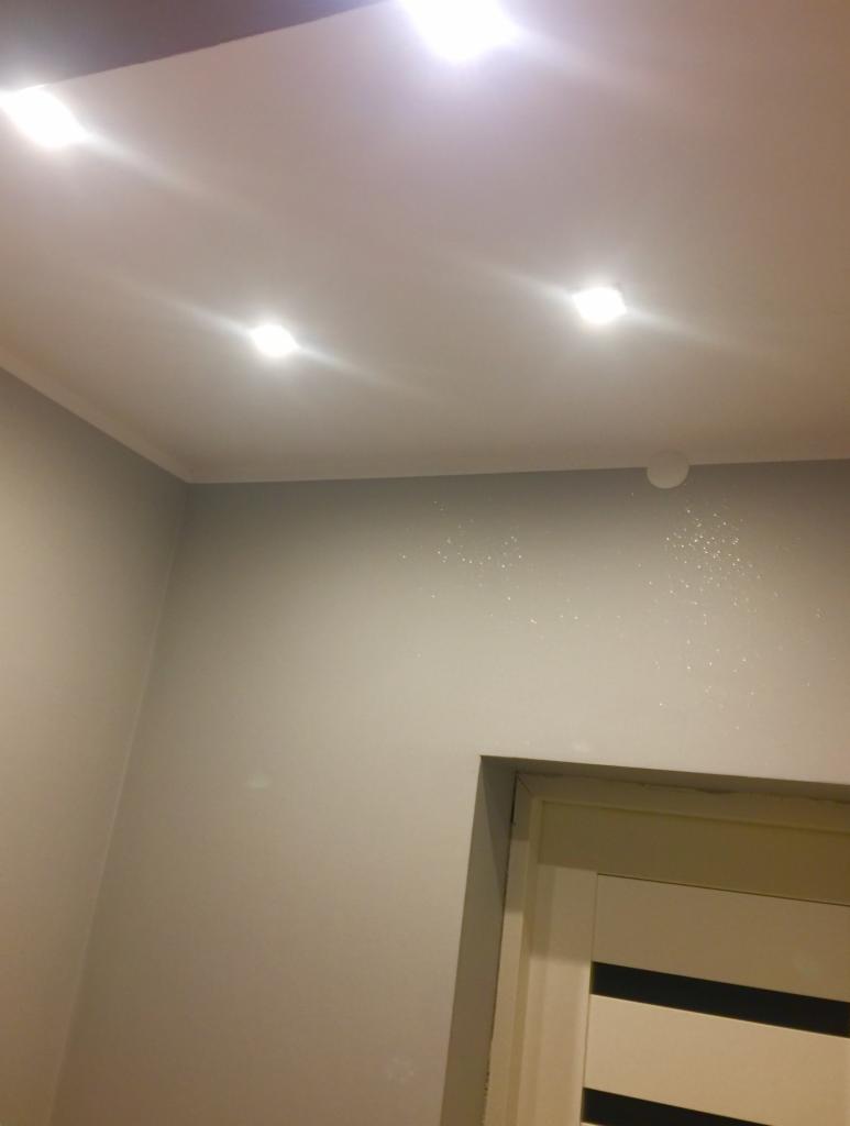 Biały sufit z czterema wpuszczonymi punktowymi lampami LED, szara ściana z delikatnym brokatem, widoczny fragment drzwi z białymi panelami.