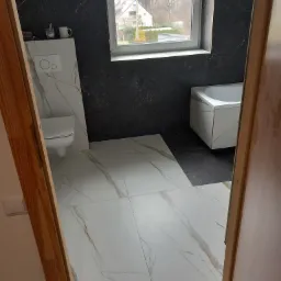 Wykończona łazienka z białymi płytkami imitującymi marmur na podłodze i ścianach, kontrastującymi ciemnymi płytkami pod oknem i przy wannie, widok zza otwartych drzwi.