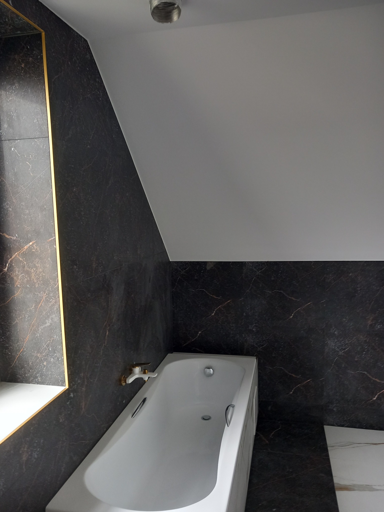 Nowoczesna łazienka z czarnymi płytkami imitującymi marmur z złotymi żyłami, biała wanna i skośny sufit.