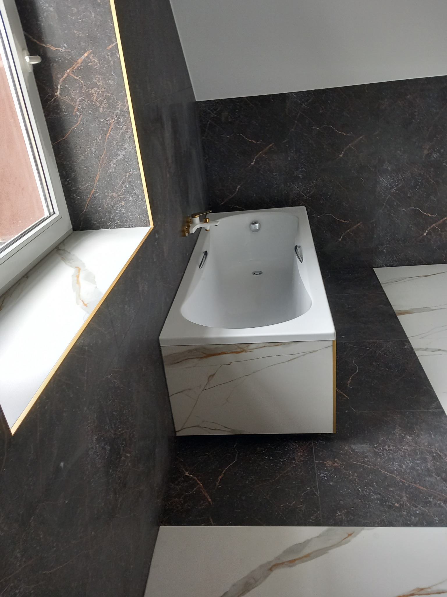 Wykończona łazienka z wanną obudowaną płytkami imitującymi marmur, czarne ściany z akcentami w kolorze złota, biały parapet z płytek z marmurowym wzorem.