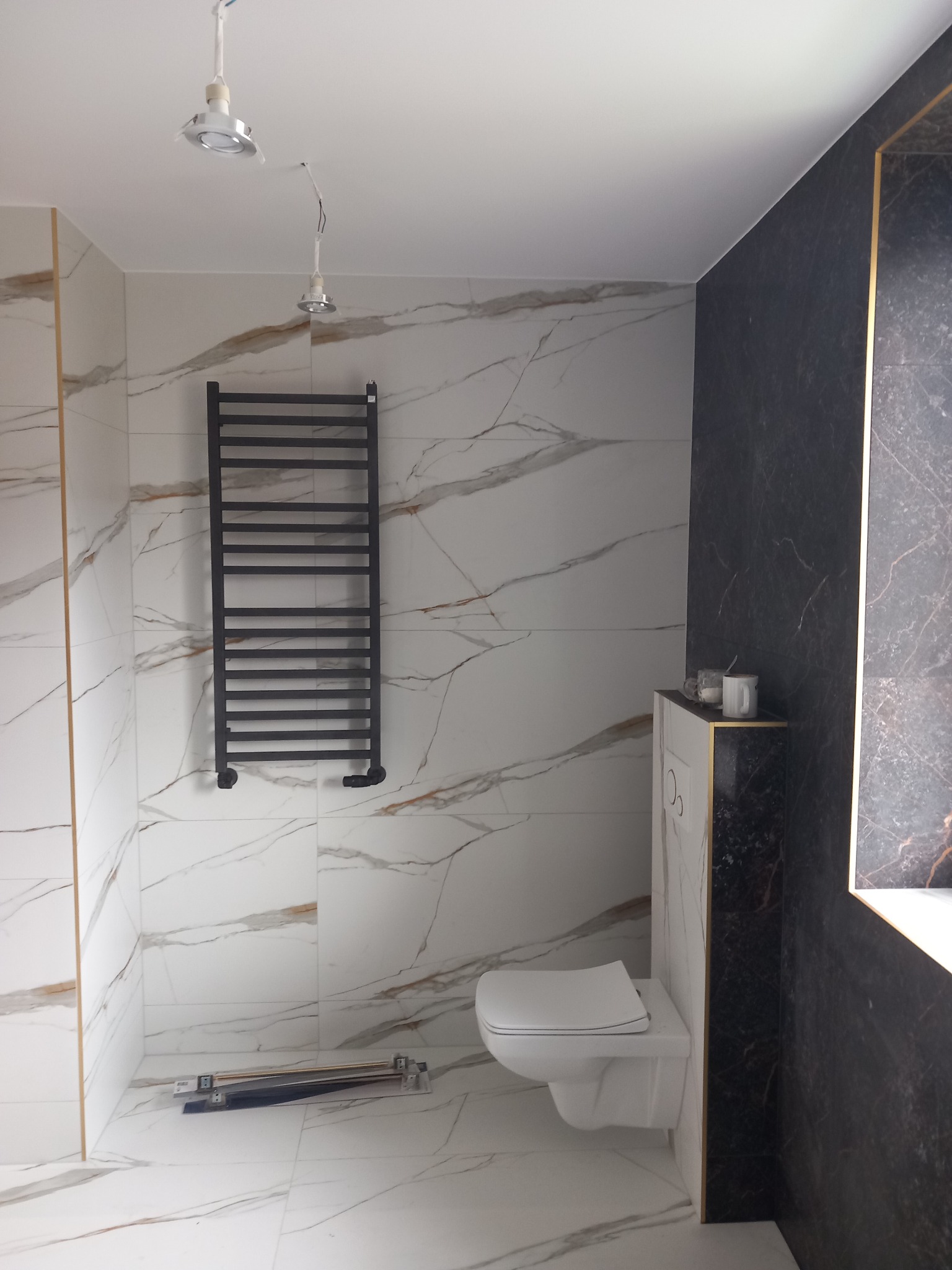 Wykończona łazienka z biało-złotymi płytkami imitującymi marmur, czarnym panelem ściennym z brokatowymi żyłkami, wiszącą toaletą i czarnym grzejnikiem drabinkowym. Na podłodze leżą narzędzia.