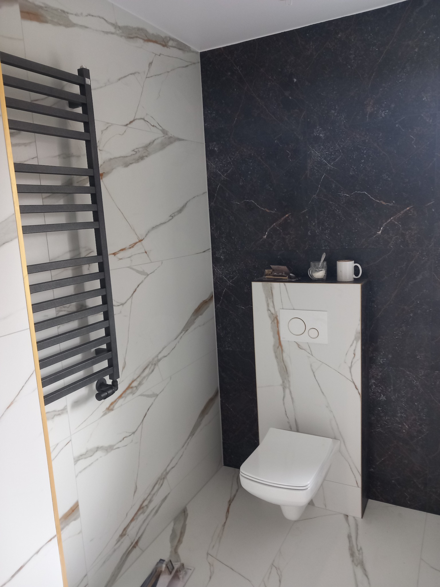 Nowoczesna łazienka z białymi i czarnymi płytkami imitującymi marmur, wisząca toaleta, czarny grzejnik drabinkowy oraz akcesoria łazienkowe na zabudowie geberit.