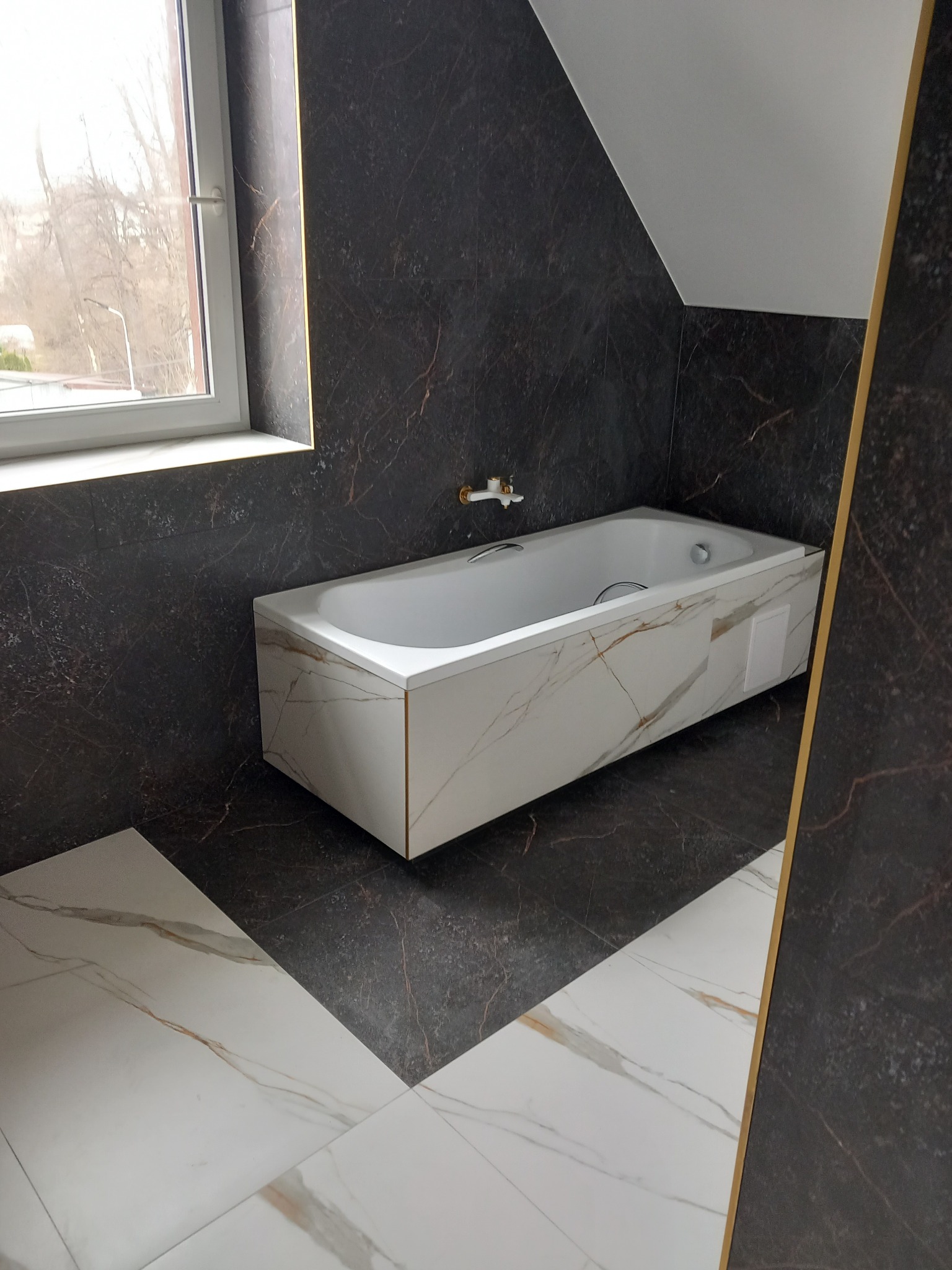 Nowoczesna łazienka z wanną obudowaną płytami imitującymi marmur, ciemne płytki ścienne z delikatnym żyłkowaniem, jasna podłoga z geometrycznym wzorem płytek, okno w tle.