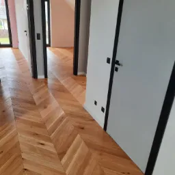 Parkiet ułożony we wzór jodełki francuskiej, z widocznymi trzema parami drzwi z czarnymi futrynami i białymi skrzydłami w nowoczesnym wnętrzu.