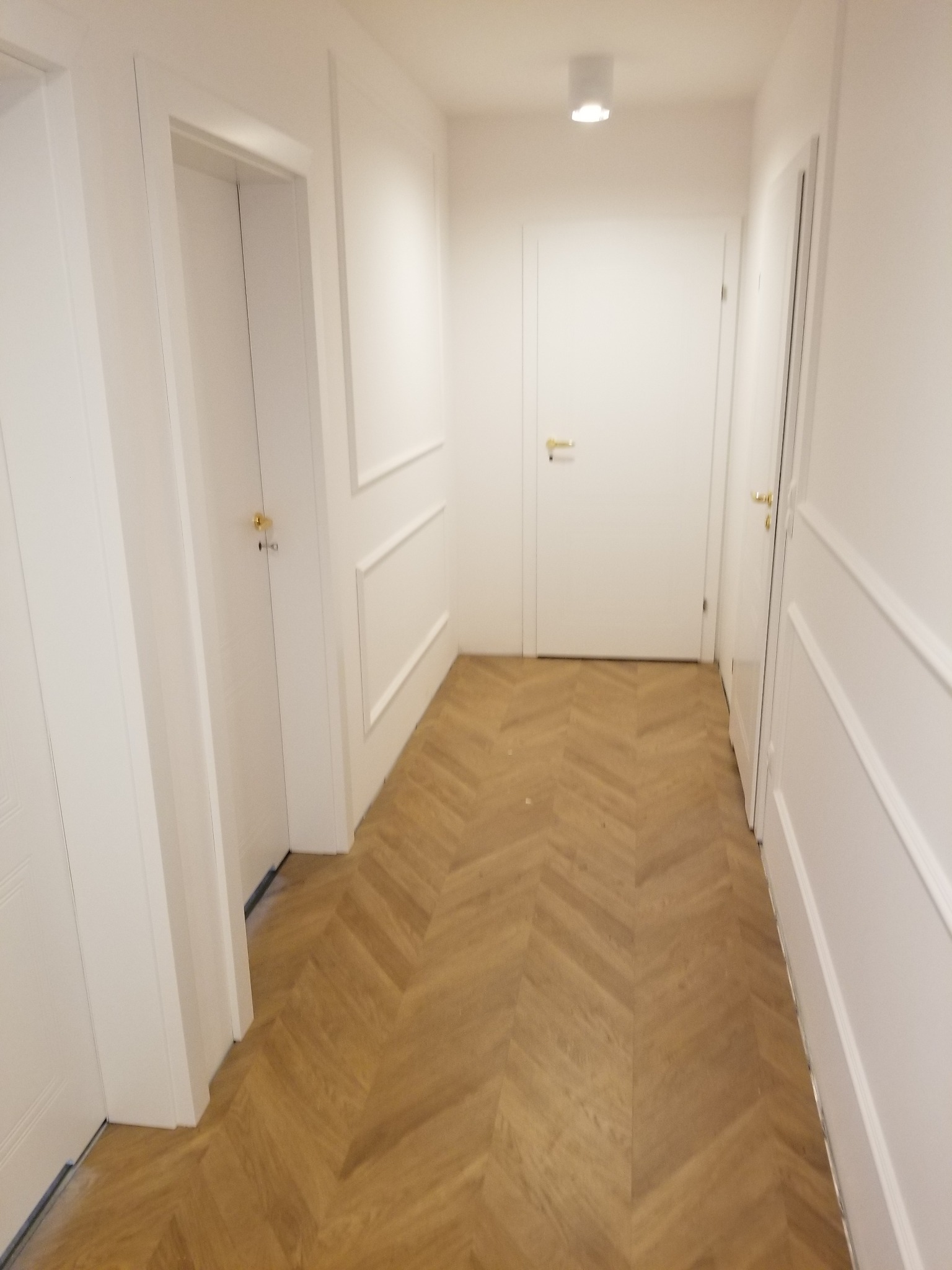Korytarz z jasnymi ścianami, białymi drzwiami z złotymi klamkami i parkietem ułożonym w jodełkę francuską, oświetlony punktowym światłem sufitowym.